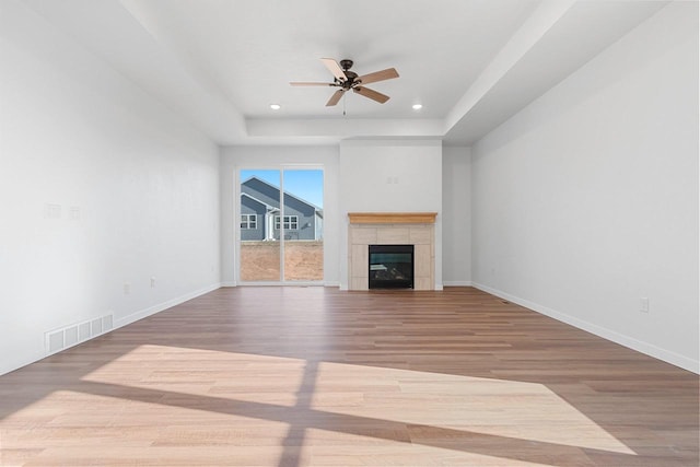 unfurnished living room with a tiled fireplace, a raised ceiling, ceiling fan, and light wood-type flooring