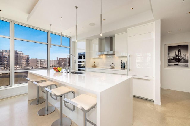 kitchen featuring a kitchen island with sink, hanging light fixtures, wall chimney range hood, and white cabinetry