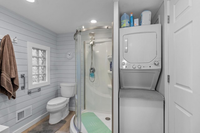 bathroom with wooden walls, hardwood / wood-style floors, a shower with door, stacked washer / dryer, and toilet