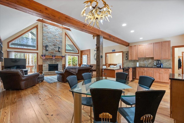 dining room with light hardwood / wood-style floors, an inviting chandelier, beam ceiling, a stone fireplace, and high vaulted ceiling