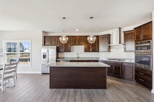 kitchen featuring dark brown cabinetry, stainless steel appliances, wood finished floors, wall chimney range hood, and a center island