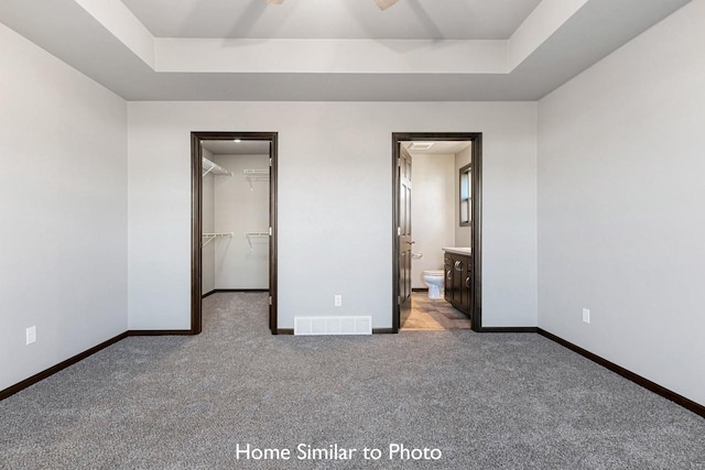 unfurnished bedroom with a closet, a raised ceiling, ensuite bath, dark carpet, and a walk in closet