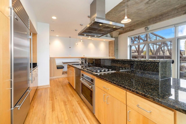 kitchen with island exhaust hood, hanging light fixtures, dark stone counters, light hardwood / wood-style floors, and stainless steel appliances