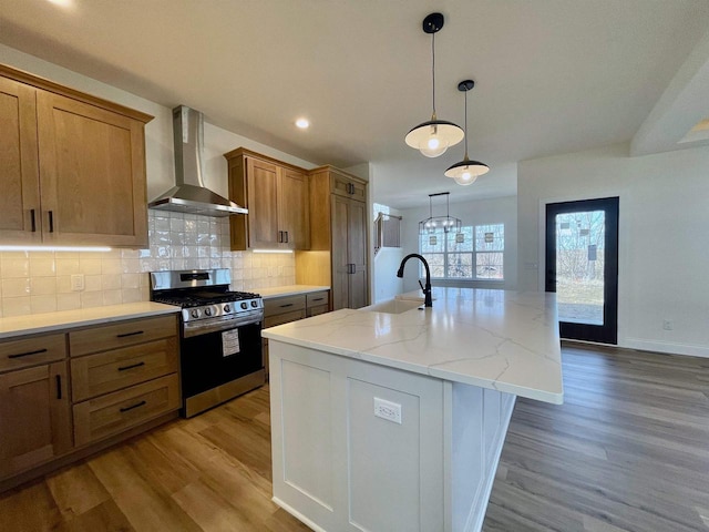 kitchen with wall chimney exhaust hood, stainless steel gas range, sink, tasteful backsplash, and a kitchen island with sink