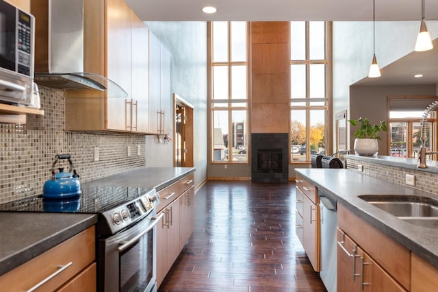 kitchen featuring wall chimney exhaust hood, decorative light fixtures, appliances with stainless steel finishes, a fireplace, and dark hardwood / wood-style flooring