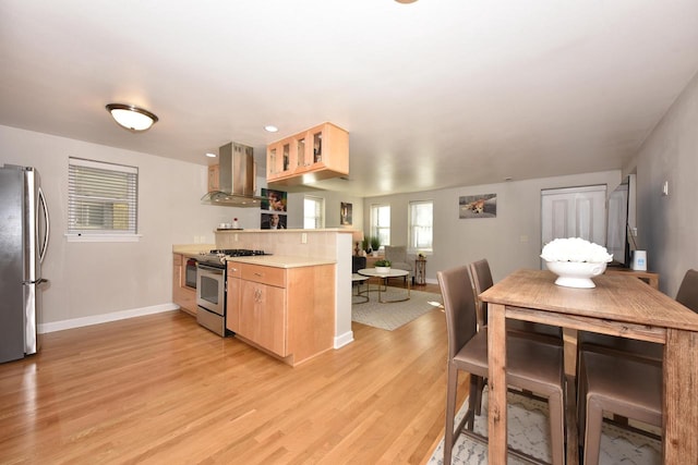 kitchen with wall chimney range hood, kitchen peninsula, stainless steel appliances, and light hardwood / wood-style floors