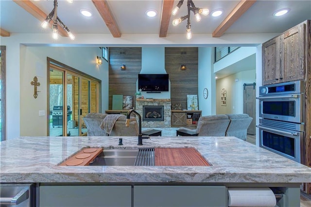 kitchen featuring light stone counters, beamed ceiling, a fireplace, sink, and stainless steel double oven