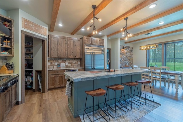 kitchen featuring beam ceiling, hanging light fixtures, appliances with stainless steel finishes, a kitchen island with sink, and light hardwood / wood-style floors