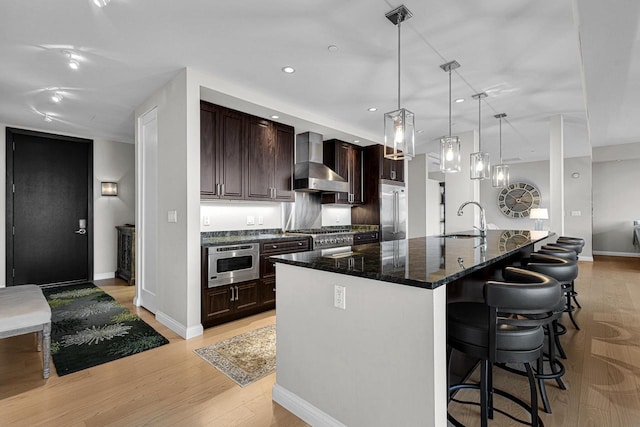 kitchen with a large island with sink, sink, light wood-type flooring, hanging light fixtures, and wall chimney exhaust hood