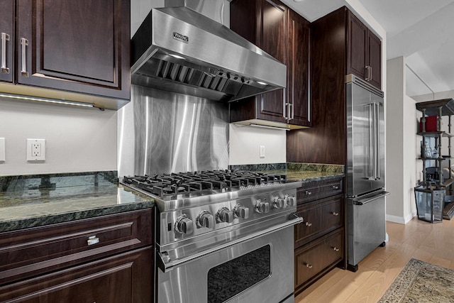 kitchen with high quality appliances, wall chimney range hood, dark stone counters, and light wood-type flooring