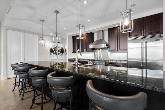 kitchen with stainless steel built in fridge, wall chimney range hood, dark stone counters, sink, and light hardwood / wood-style floors