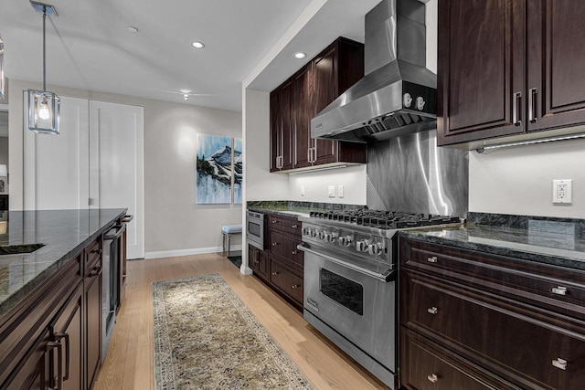 kitchen featuring wall chimney exhaust hood, dark stone countertops, decorative light fixtures, appliances with stainless steel finishes, and light hardwood / wood-style floors