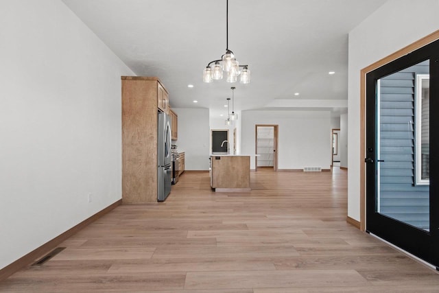 kitchen featuring light wood-type flooring, stainless steel fridge, decorative light fixtures, light brown cabinets, and a kitchen island with sink