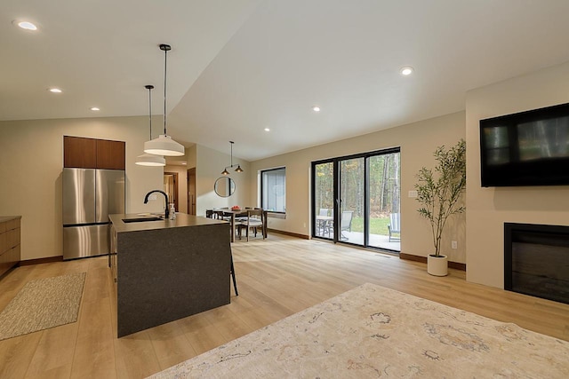 kitchen with an island with sink, light hardwood / wood-style flooring, sink, stainless steel fridge, and vaulted ceiling