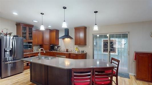 kitchen with wall chimney range hood, sink, light wood-type flooring, an island with sink, and appliances with stainless steel finishes