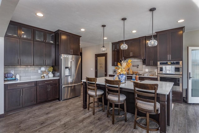 kitchen with a center island with sink, stainless steel appliances, backsplash, pendant lighting, and dark wood-type flooring