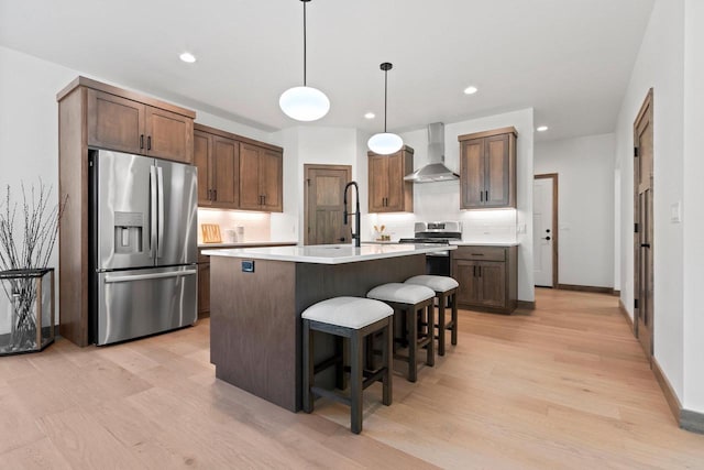 kitchen with hanging light fixtures, light hardwood / wood-style floors, stainless steel appliances, wall chimney exhaust hood, and a kitchen island with sink