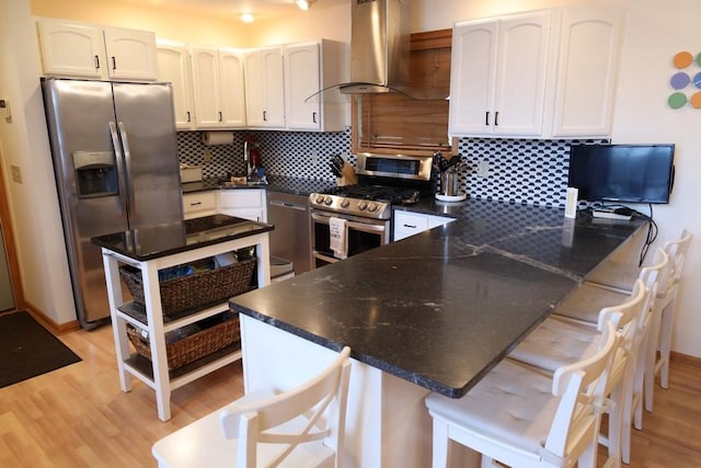 kitchen with wall chimney range hood, appliances with stainless steel finishes, a breakfast bar, white cabinets, and light hardwood / wood-style flooring