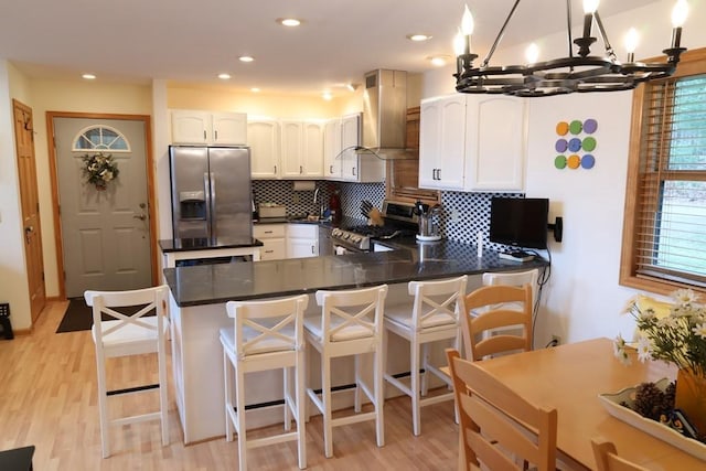 kitchen featuring stainless steel appliances, white cabinetry, kitchen peninsula, wall chimney exhaust hood, and a breakfast bar area