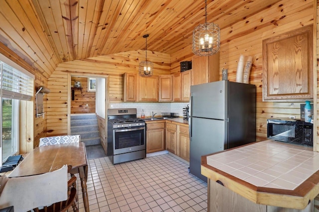 kitchen featuring tile counters, vaulted ceiling, hanging light fixtures, wooden walls, and appliances with stainless steel finishes