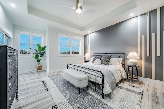 bedroom featuring multiple windows, light hardwood / wood-style flooring, ceiling fan, and a tray ceiling