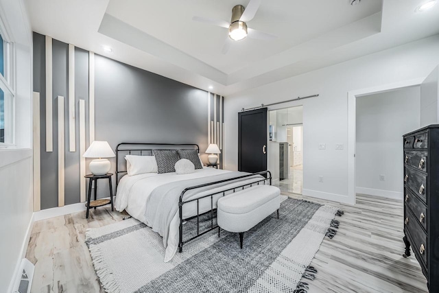 bedroom with light hardwood / wood-style floors, a barn door, ceiling fan, and a raised ceiling