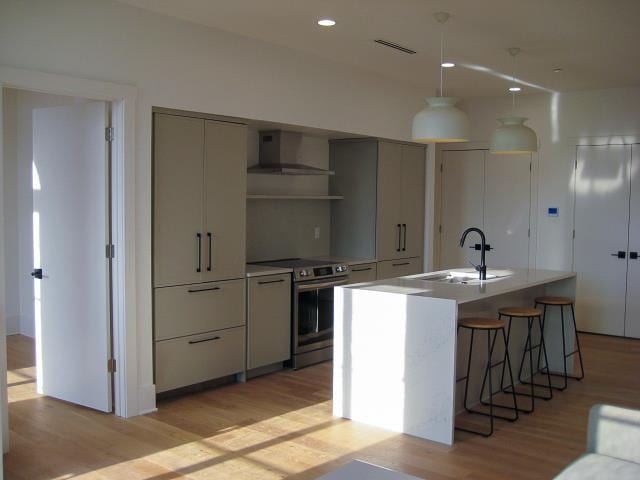 kitchen featuring stainless steel range with electric stovetop, a center island with sink, light wood-type flooring, sink, and wall chimney exhaust hood