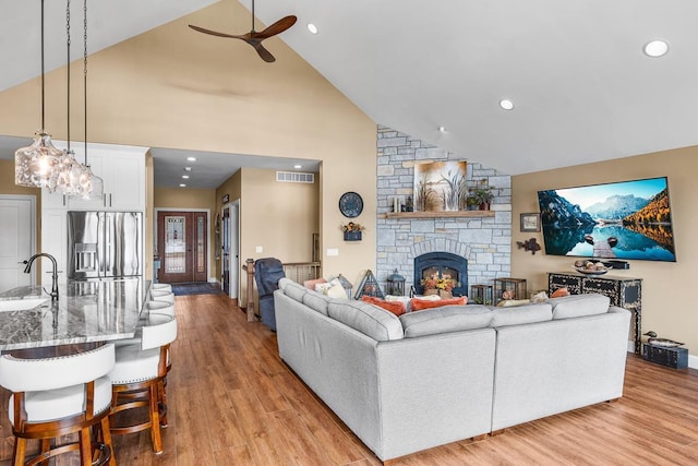 living room featuring ceiling fan, sink, high vaulted ceiling, light hardwood / wood-style floors, and a stone fireplace