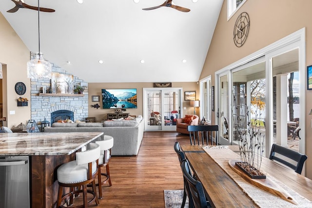 dining space featuring ceiling fan, french doors, dark wood-type flooring, a stone fireplace, and high vaulted ceiling