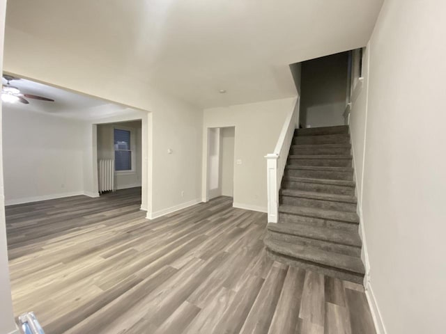 unfurnished living room with wood finished floors, ceiling fan, stairs, baseboards, and radiator heating unit