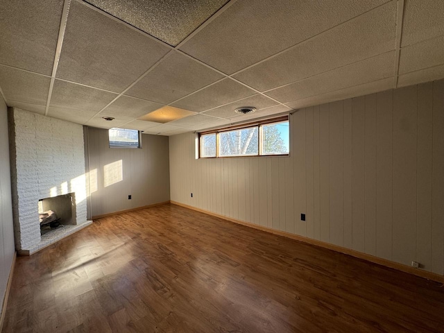 basement with a drop ceiling, wood walls, a stone fireplace, and wood-type flooring