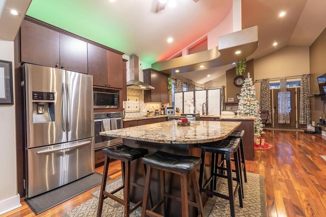 kitchen with wall chimney range hood, decorative backsplash, appliances with stainless steel finishes, dark brown cabinets, and a kitchen island