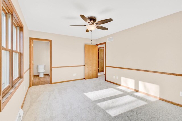 unfurnished bedroom featuring ceiling fan, light colored carpet, multiple windows, and ensuite bath