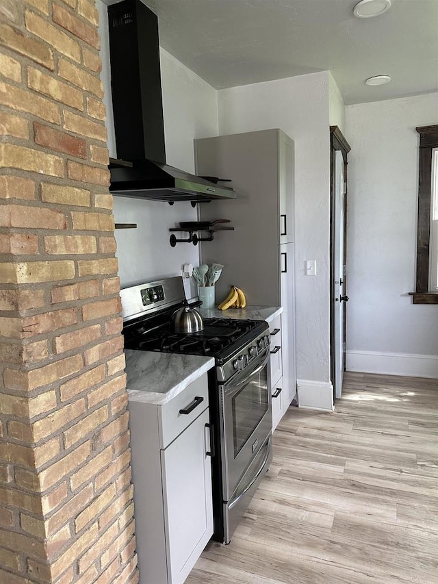 kitchen featuring light wood-style flooring, white cabinets, stainless steel gas stove, wall chimney range hood, and baseboards