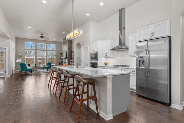 kitchen with dark wood-type flooring, wall chimney range hood, a center island with sink, white cabinets, and appliances with stainless steel finishes