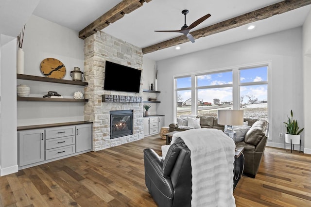 living room featuring ceiling fan, a stone fireplace, beam ceiling, and hardwood / wood-style floors