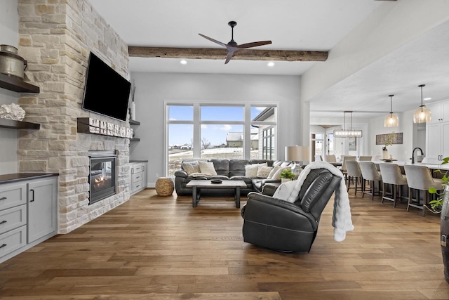 living room featuring hardwood / wood-style flooring, a stone fireplace, ceiling fan with notable chandelier, and beam ceiling