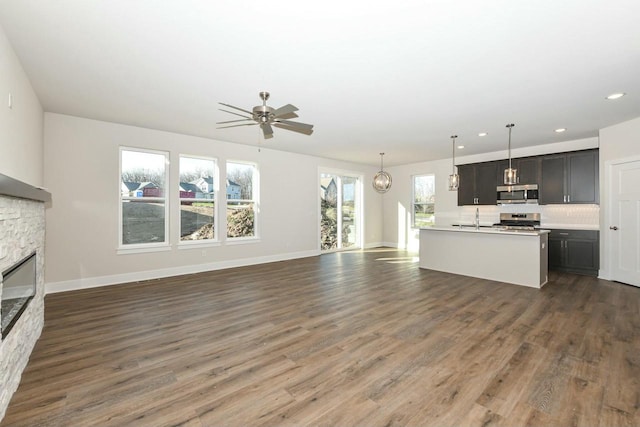 kitchen with ceiling fan, dark wood-type flooring, stainless steel appliances, a kitchen island with sink, and a fireplace