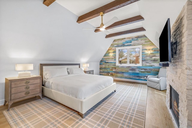 bedroom featuring hardwood / wood-style floors, lofted ceiling with beams, a stone fireplace, and ceiling fan