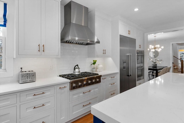 kitchen with backsplash, wall chimney exhaust hood, appliances with stainless steel finishes, decorative light fixtures, and white cabinetry