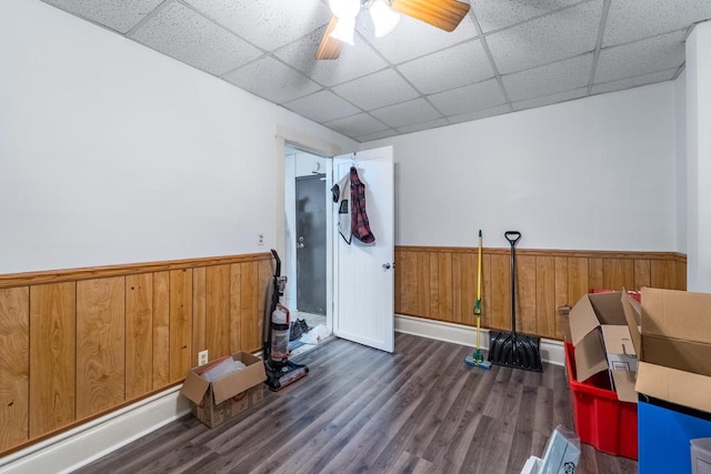 exercise room with a paneled ceiling, wooden walls, ceiling fan, and dark wood-type flooring