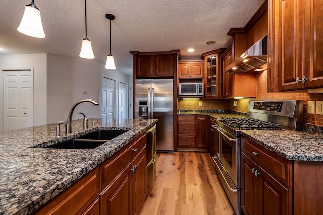 kitchen with light wood-type flooring, wall chimney exhaust hood, stainless steel appliances, sink, and hanging light fixtures