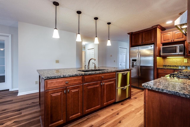 kitchen with appliances with stainless steel finishes, sink, wood-type flooring, pendant lighting, and an island with sink