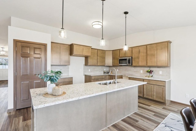 kitchen featuring stainless steel microwave, decorative backsplash, a kitchen island with sink, a sink, and wood finished floors