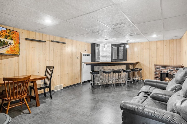 living room with a paneled ceiling, a brick fireplace, wooden walls, and bar area