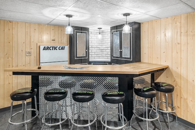 bar with light brown cabinets, a drop ceiling, hanging light fixtures, and wooden walls