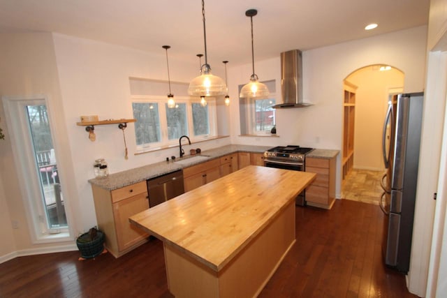 kitchen with wall chimney exhaust hood, appliances with stainless steel finishes, decorative light fixtures, a kitchen island, and dark hardwood / wood-style flooring