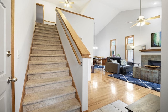 stairs with ceiling fan, wood-type flooring, high vaulted ceiling, and a tiled fireplace