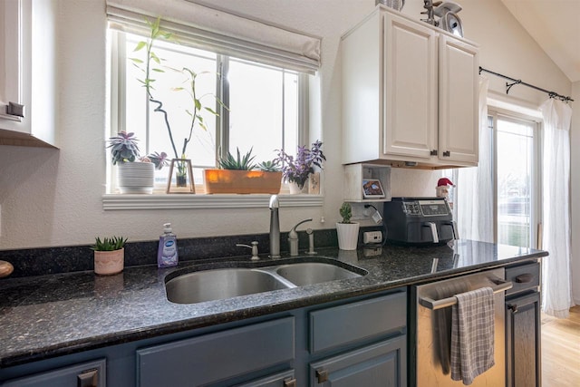 kitchen featuring stainless steel dishwasher, dark stone counters, vaulted ceiling, sink, and white cabinets