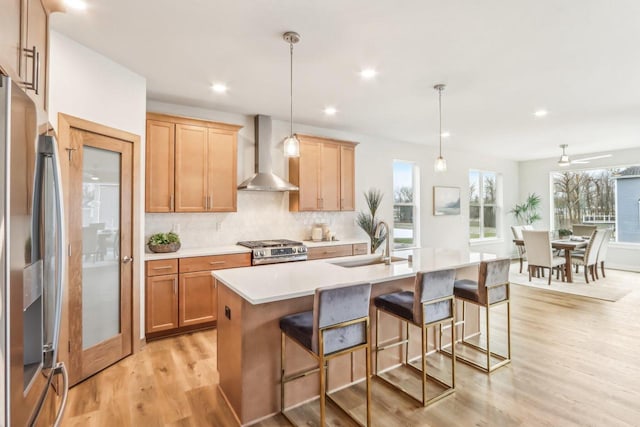 kitchen with a kitchen island with sink, wall chimney range hood, sink, hanging light fixtures, and appliances with stainless steel finishes